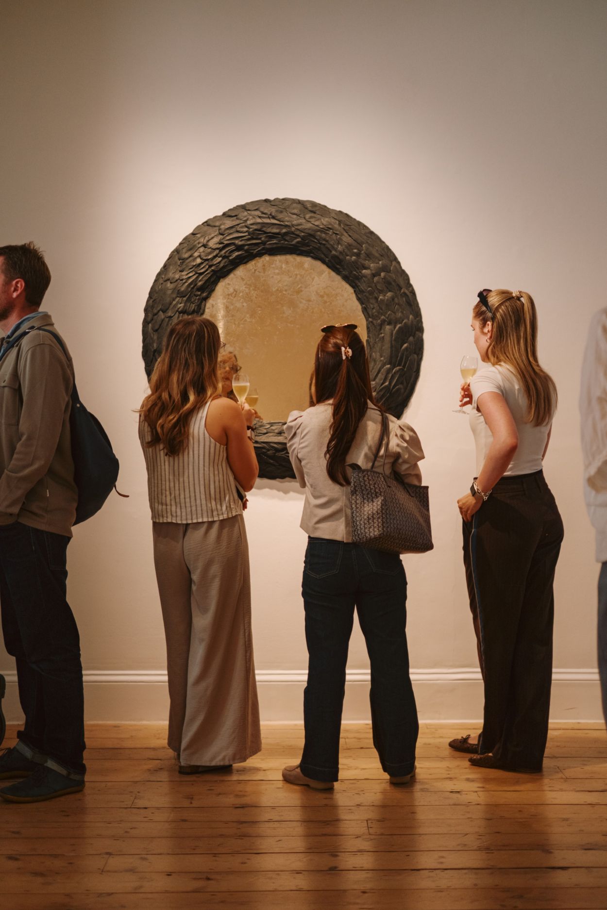 A crowd at a gallery looking at a large, round mirror of sculpted feathers and gilding.